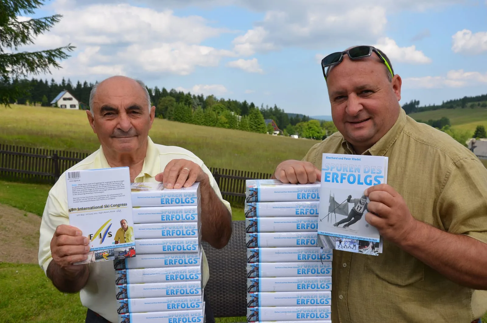 Eberhard und Peter Riedel halten je ein Exemplar von „Spuren des Erfolgs“ vor einem Stapel des Buches in die Kamera