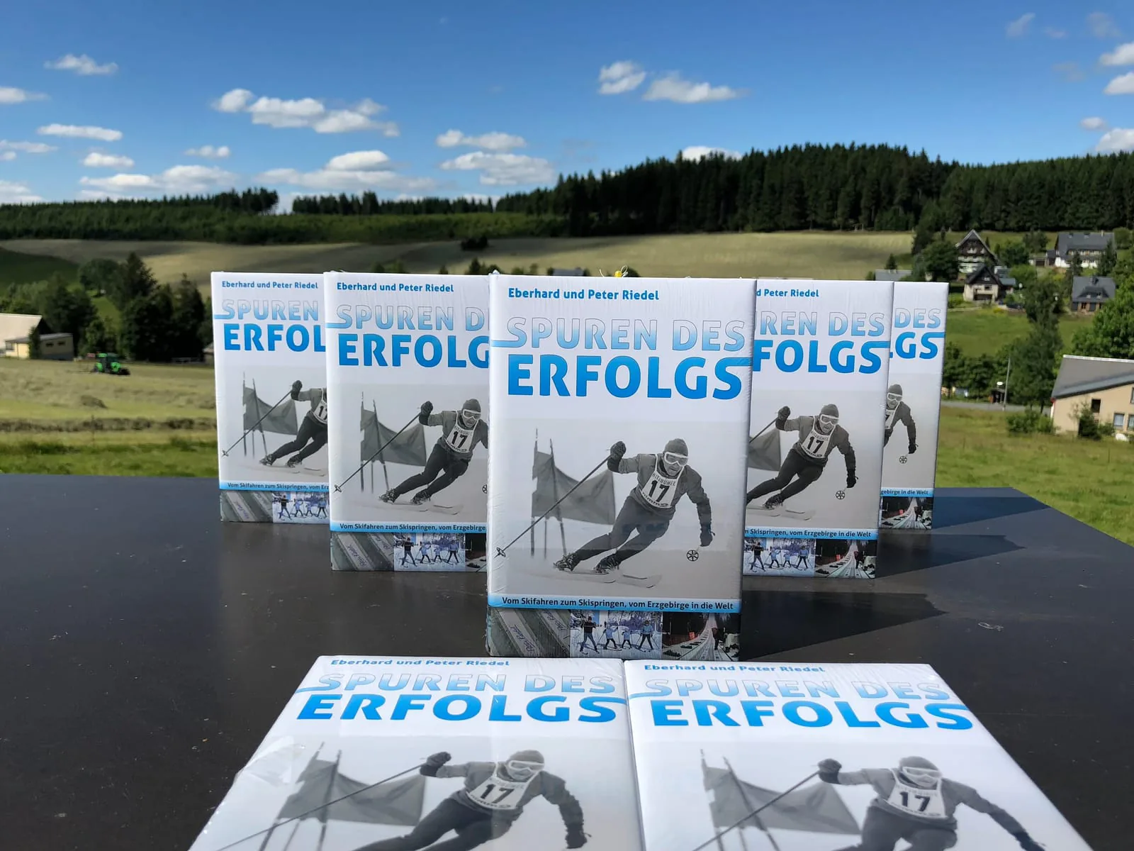 Mehrere eingeschweißte Exemplare von „Spuren des Erfolgs“ auf einem Tisch, dahinter die Wiesen und Wälder des Erzgebirges