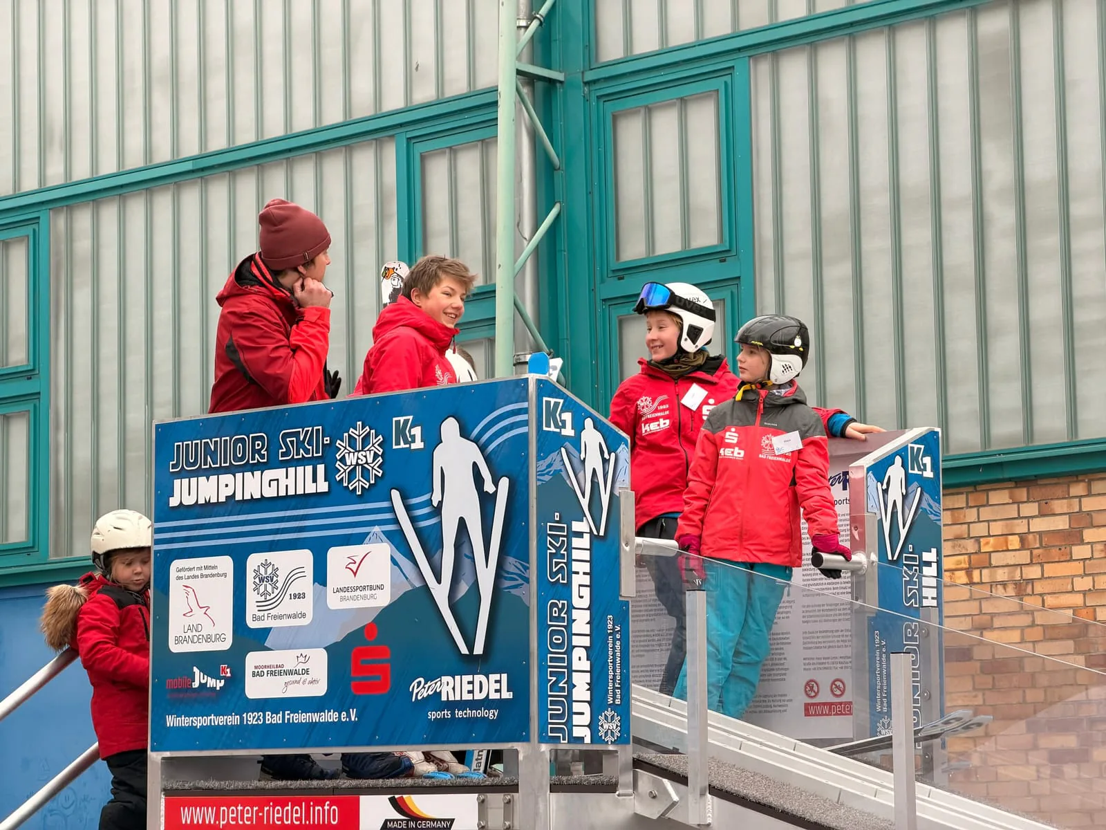 Kinder und Betreuer auf der Startplattform der MOBILE JUMP hinter dem Banner Junior Ski Jumping Hill