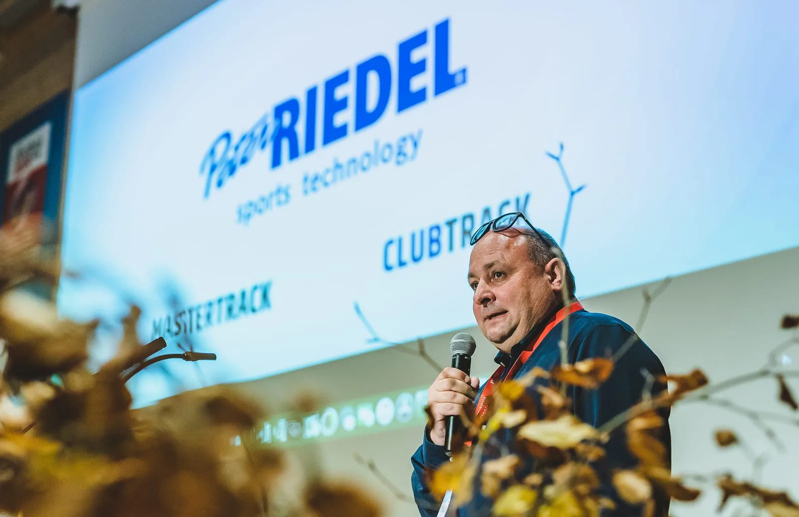 Peter Riedel spricht mit Mikrofon vor einer Leinwand mit dem Schriftzug Peter Riedel Sports Technology, MASTERTRACK und CLUBTRACK